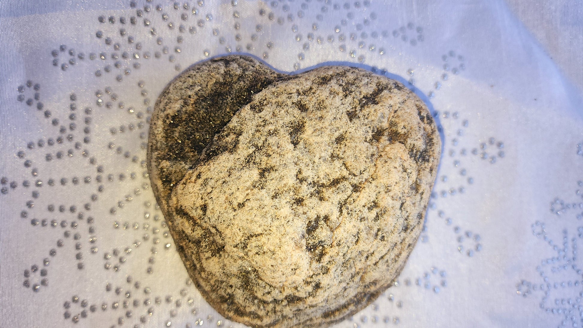 Rounded, speckled heart-shaped stone in shades of tan, black, and gold, shown resting on white snowflake fabric and cradled in hand.